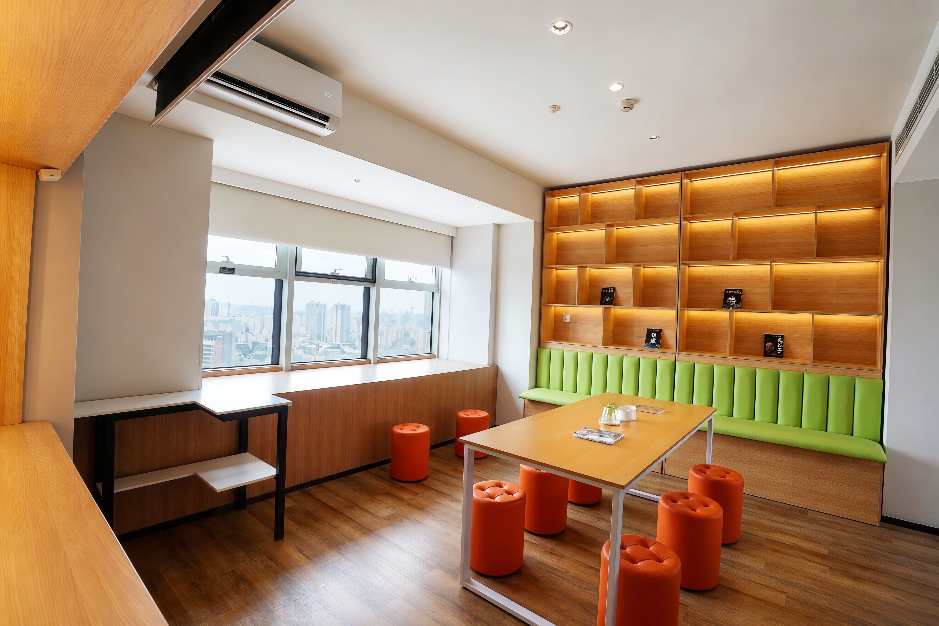 Un interior moderno con un escritorio de madera, taburetes naranjas, un banco acolchado verde y vistas a la ciudad a través de amplios ventanales.