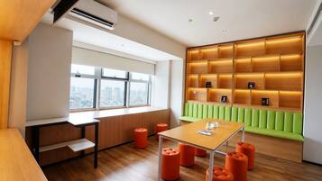 A modern interior with a wooden desk, orange stools, a green cushioned bench, and a city view through large windows.