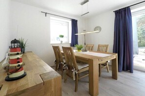 A dining area with a wooden table, four chairs, a clock on the wall, and a window with curtains. - Holiday home with 2 bedrooms (Dierhagen)