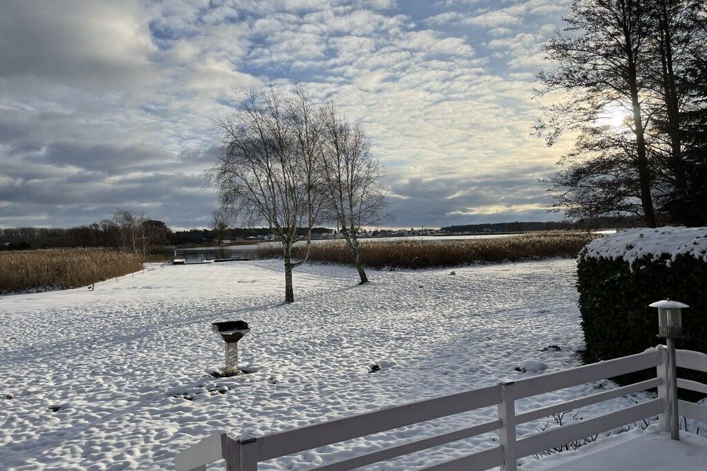 Eine verschneite Landschaft mit einem weißen Zaun, einem Laternenpfahl und kahl stehenden Bäumen.