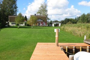 Ett grönområde med en träbrygga som sträcker sig ut i vattnet, omgivet av träd och ett hus i bakgrunden.