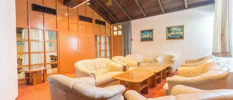 Une pièce avec un plafond haut, des poutres en bois, une grande fenêtre avec des rideaux et un coin salon comprenant des canapés et une table basse.
