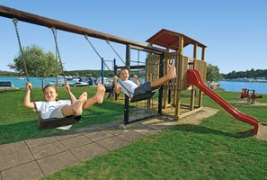 A playground with swings and a slide, located near a body of water with boats in the background. - Apartments Laguna Galijot (Porec)