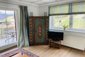 Une pièce avec un téléviseur posé sur un meuble, une fenêtre donnant sur l’extérieur, un meuble orné de motifs décoratifs et une table avec une chaise à l’extérieur.