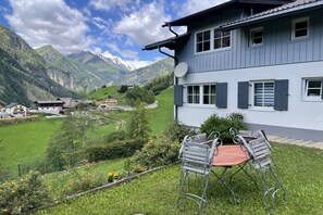 Un bâtiment à deux étages, doté d’une antenne parabolique, d’un espace repas avec des chaises en métal, et offrant une vue sur une vallée avec des montagnes en arrière-plan.