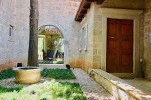 A stone building with a wooden door, a stone bench, and a small garden area with a tree and a birdbath. - Villa Tereza (Konavle)