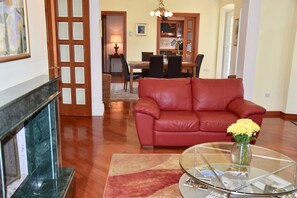 Una sala de estar con un sofá de cuero rojo, una mesa de centro de vidrio con papeles, una chimenea y un área de comedor al fondo.