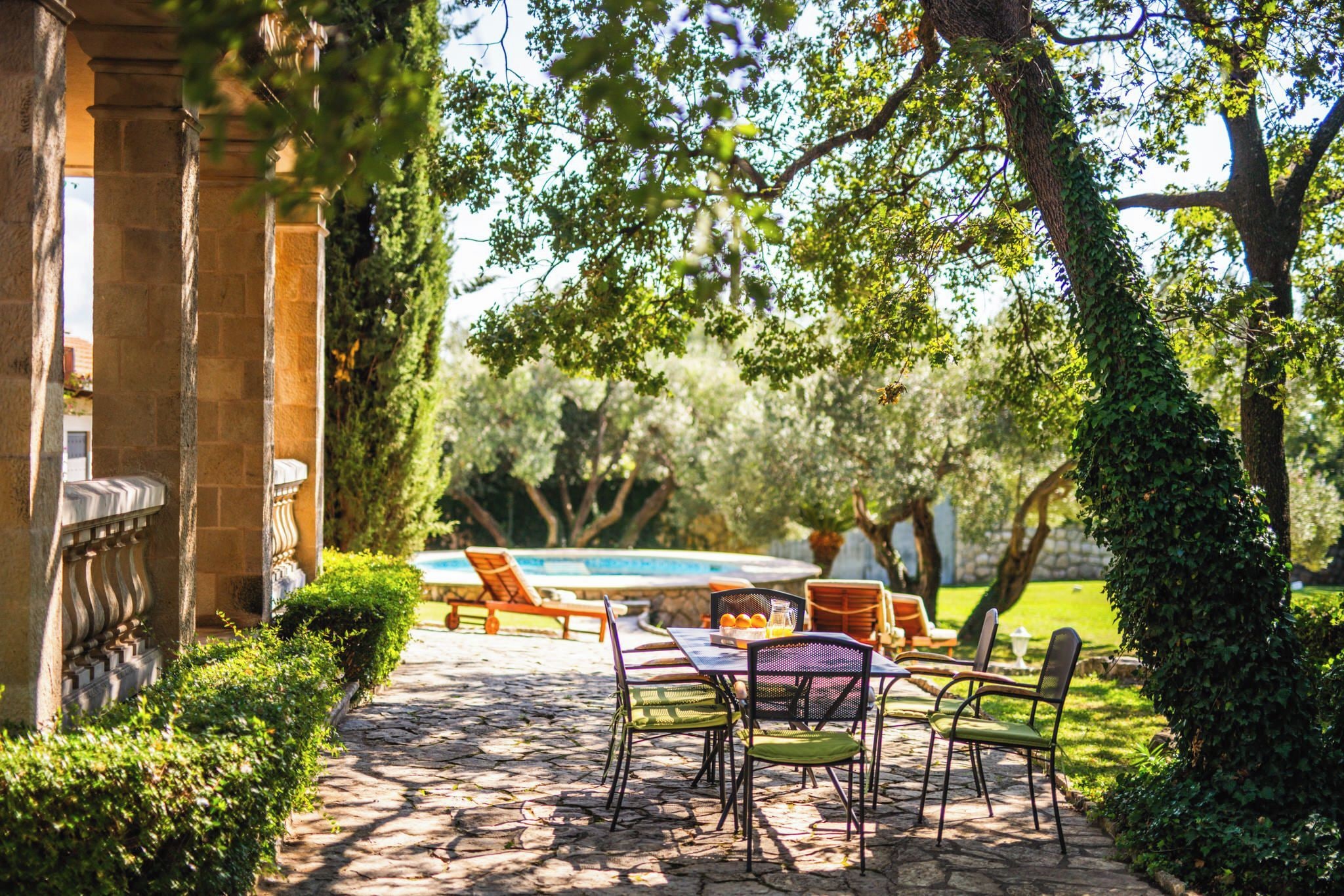 A patio area with stone flooring, outdoor seating, and a swimming pool in the background.
