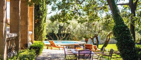 A patio area with stone flooring, outdoor seating, and a swimming pool in the background.
