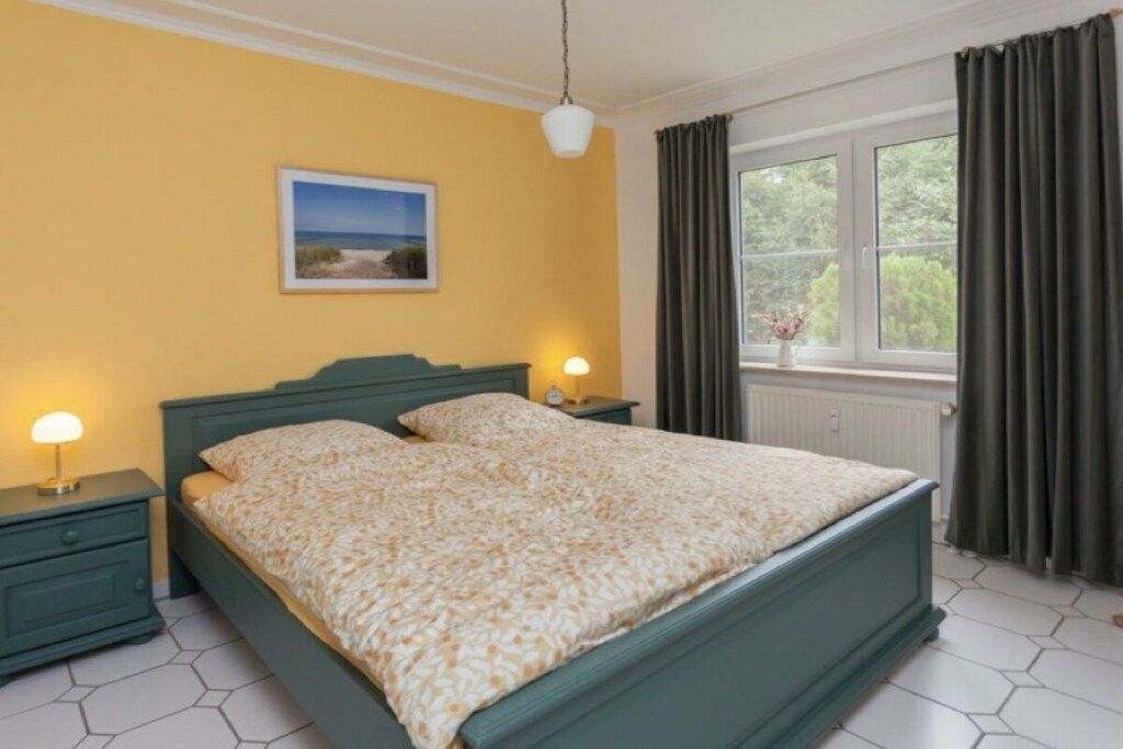 A bedroom with a bed, two bedside tables, a night lamp, a window with curtains, and a wall-mounted picture.