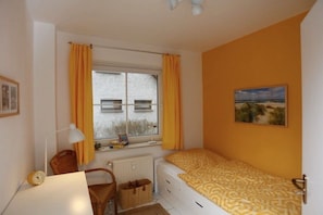 Ein Schlafzimmer mit Bett, Schreibtisch, Stuhl, einem Fenster mit Vorhängen, einem Heizkörper und einem Bild an der Wand.