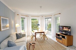 Ein Wohnzimmer mit einer Couch, einem Esstisch mit Stühlen, einer TV-Konsole und großen Fenstern.