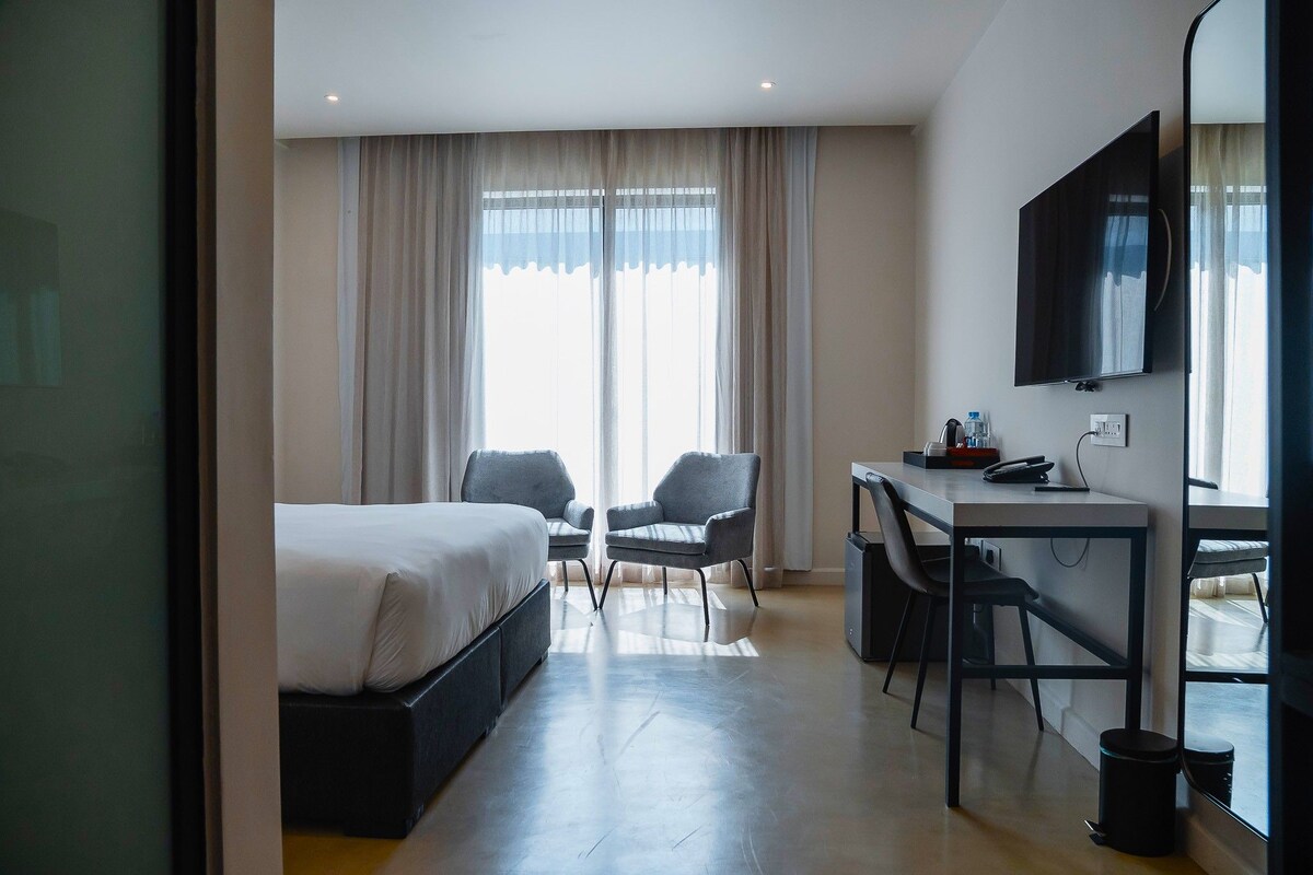 Habitació d'hotel amb un llit, dues cadires, un escriptori, una televisió i una finestra amb cortines.