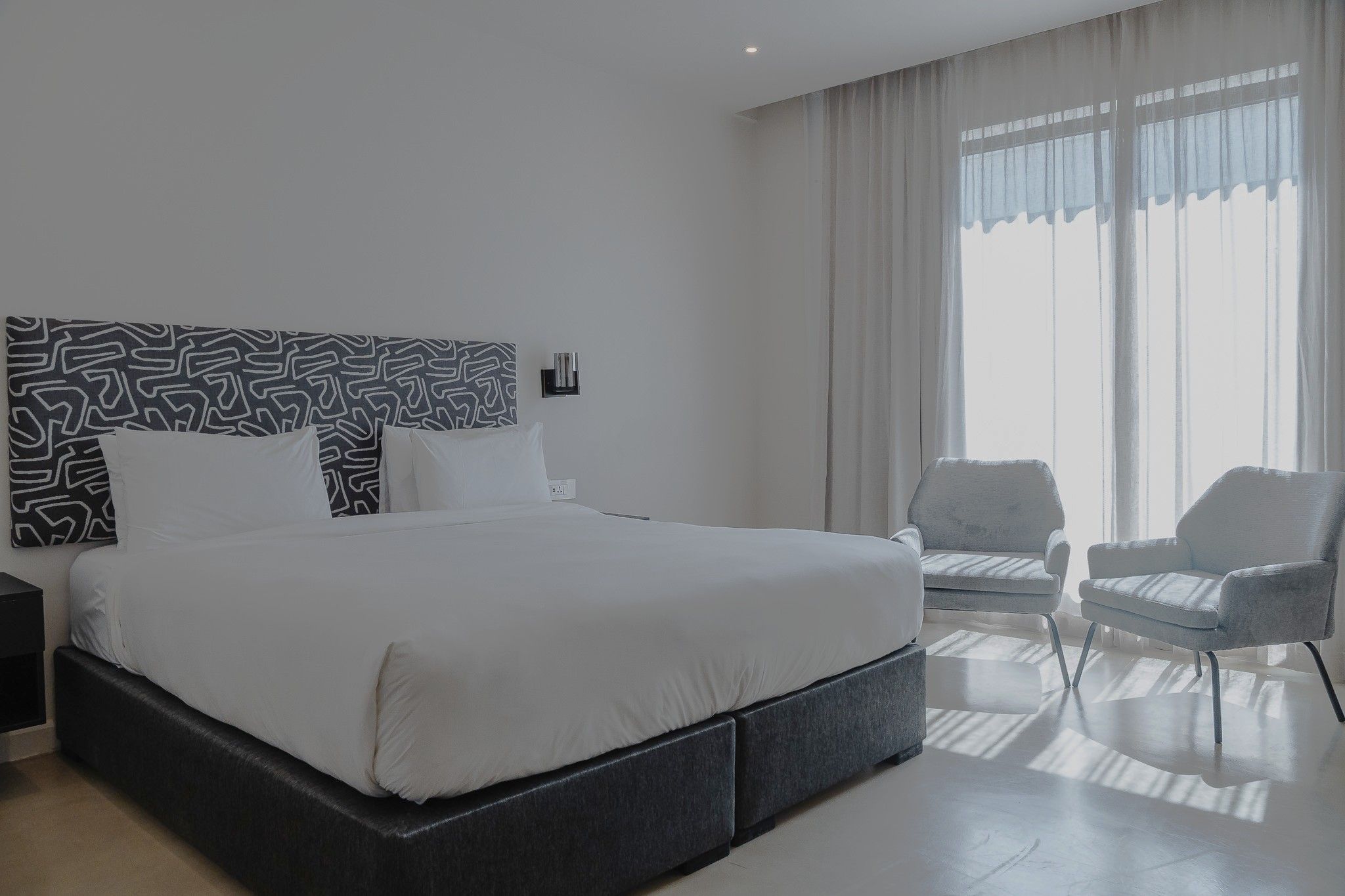 Ein modernes Hotelzimmer mit einem großen Bett, einem Nachttisch, einem Sessel und einem Fenster mit Vorhängen.