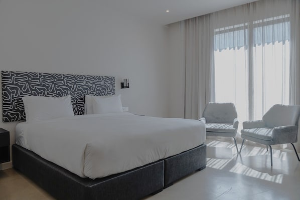Una habitación de hotel moderna con una cama grande, una mesita de noche, una silla y una ventana con cortinas.