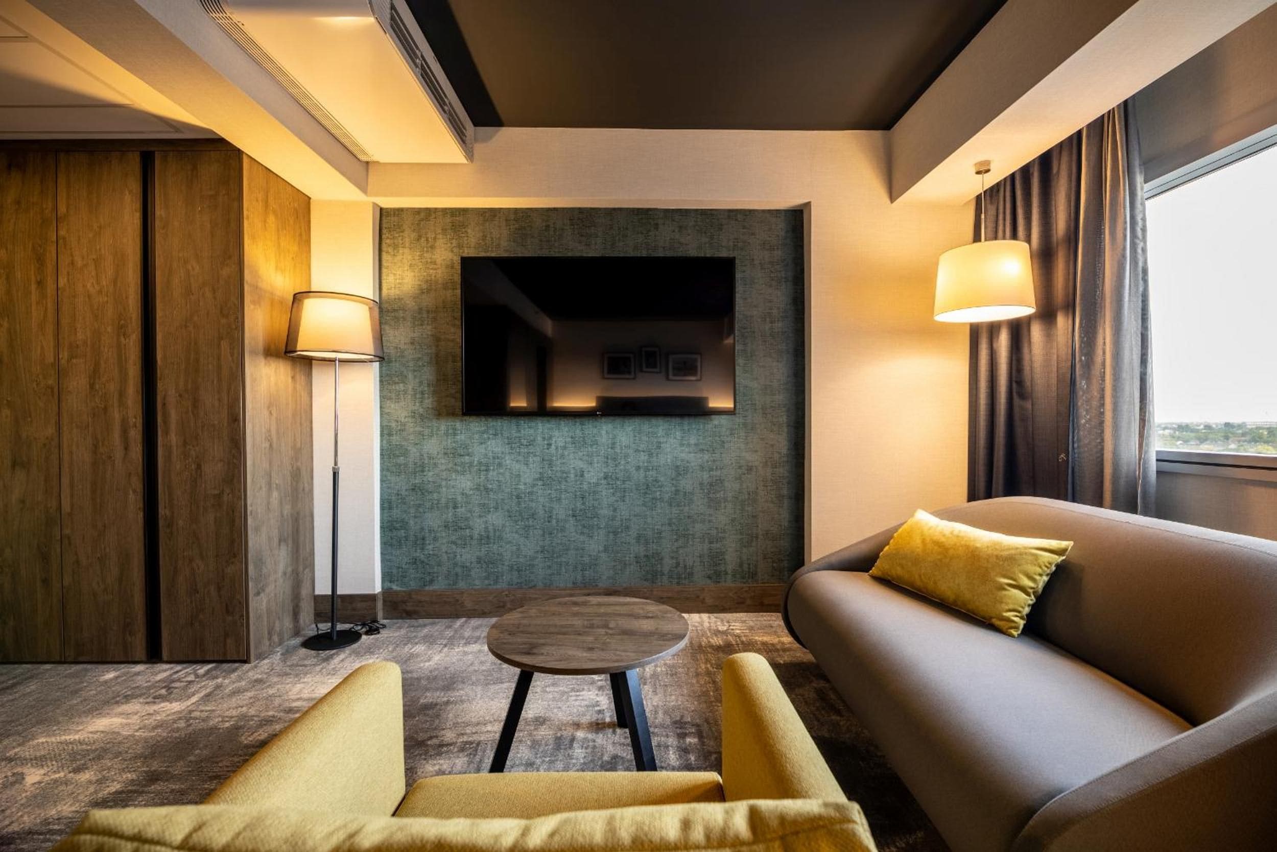 Ein modernes Hotelzimmer mit einer Couch, Sesseln, einem kleinen runden Tisch und einem an der Wand befestigten Fernseher.