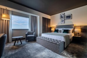 A modern hotel room with a large bed, a seating area with a chair and a small table, a window with curtains, and a wall with framed pictures. - Osijek (Osijek)
