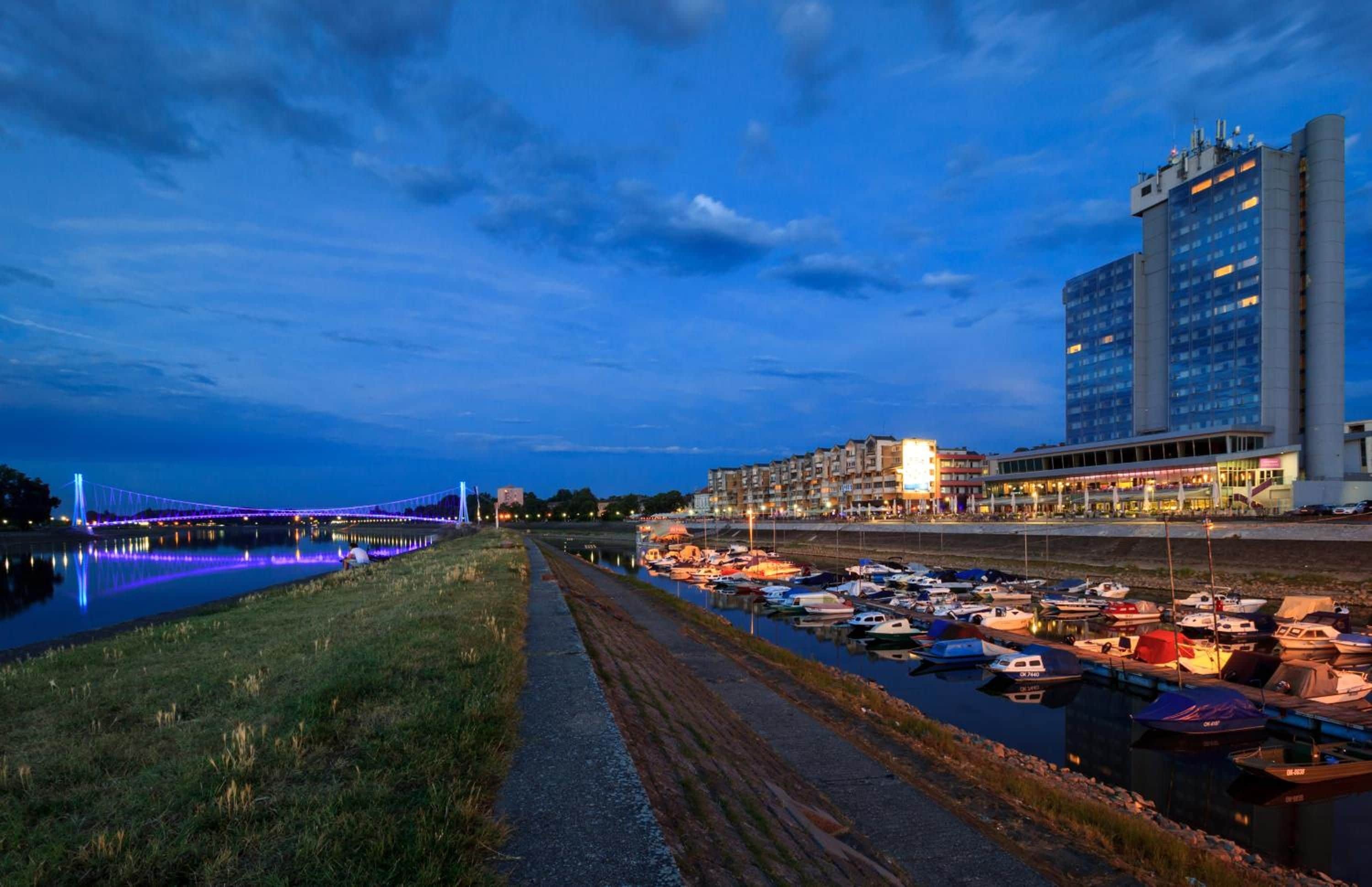 Ein Yachthafen am Wasser mit vertäuten Booten, einem Fußweg und einer Brücke mit beleuchteten Lichtern.