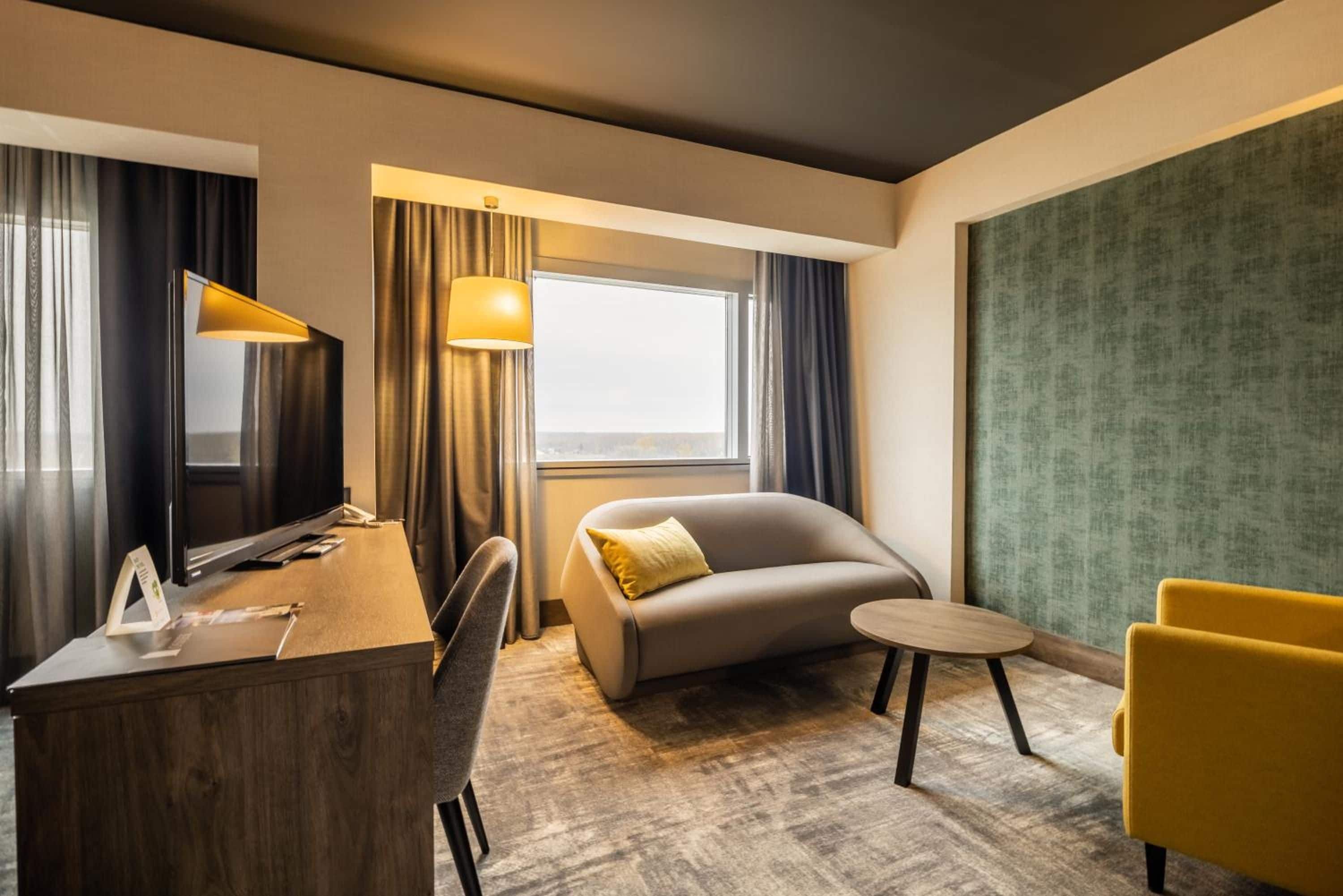 Ein Hotelzimmer mit Schreibtisch, einer Couch, einem kleinen runden Tisch und einem gelben Sessel.