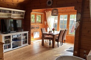 A wooden cabin interior with a dining table, chairs, a bookshelf, and a television. - Holiday home with 2 bedrooms (Ahrenshoop)