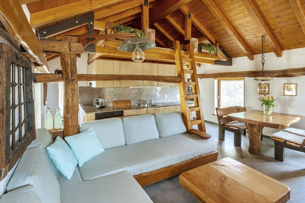 A rustic interior with a wooden ceiling, a built-in kitchen, a sofa with cushions, and a dining table with benches.