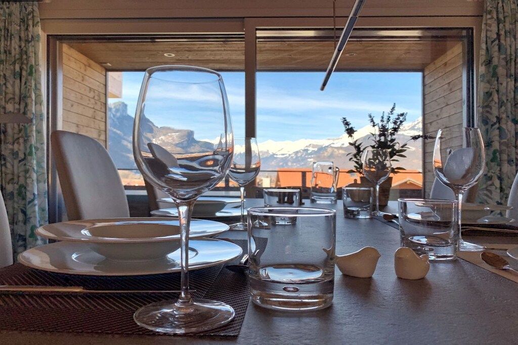 Une table à manger dressée avec des assiettes, des verres et un vase rempli de fleurs, offrant une vue sur les montagnes à travers de grandes fenêtres.