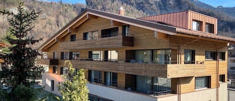 A multi-story building with balconies, large windows, and a mountainous backdrop.