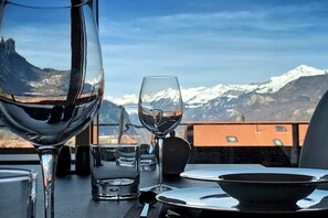 Une table à manger avec un verre à vin, un verre à eau et un bol, le tout préparé pour un repas. La table est placée à l’extérieur, avec une vue sur les montagnes en arrière-plan.