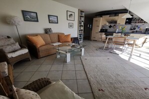 Ein Wohnzimmer mit einer Couch, einem Couchtisch und einem Essbereich mit Stühlen. Im Hintergrund sind ein Bücherregal und eine Küche erkennbar.