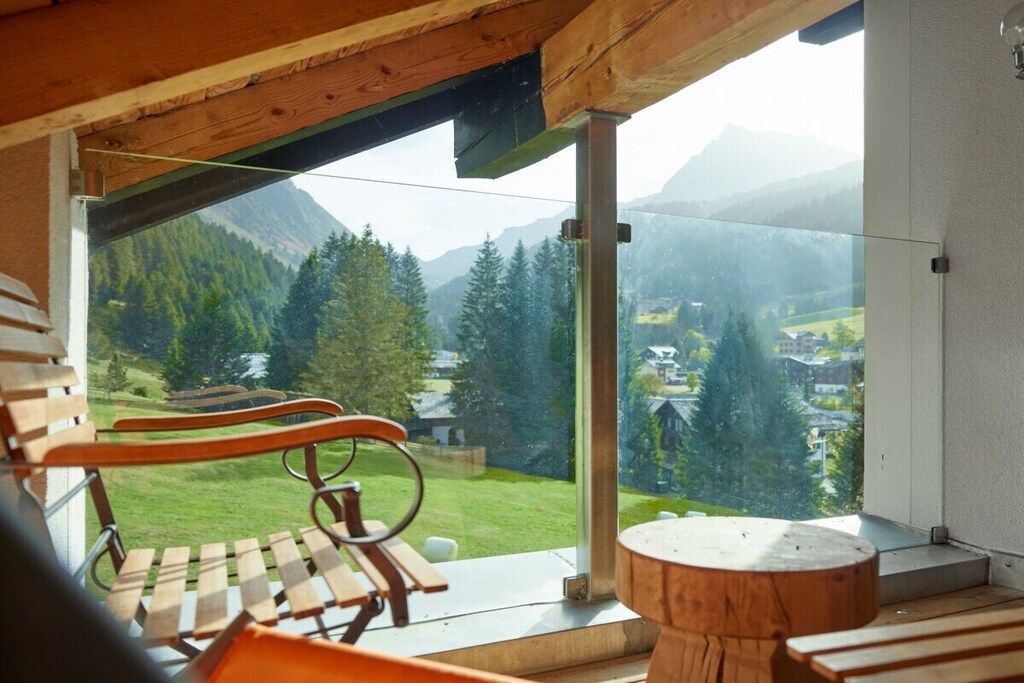 Una terraza de madera con un banco y una mesa redonda, con vistas a un paisaje montañoso con árboles y casas.