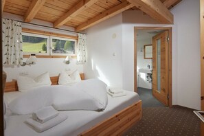 A bedroom with a wooden bed, white linens, a window with curtains, and a view of the outdoors. - 5405 Elfriede in the Alpenhaus Montafon (Bludenz)