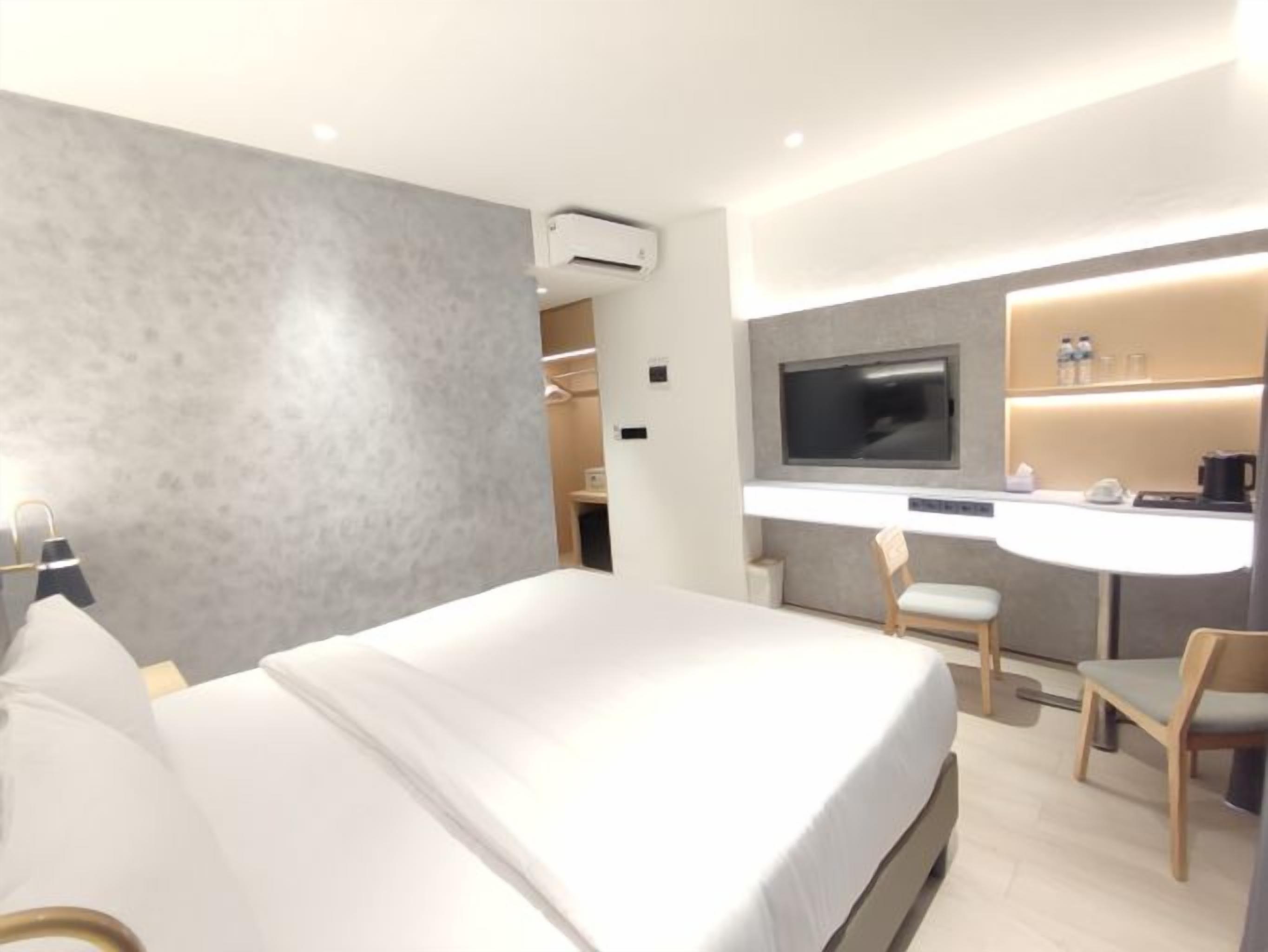 A modern hotel room with a large bed, a desk with a chair, a television, and a minibar.