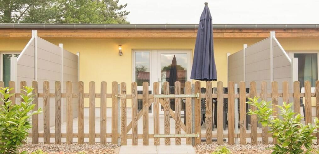 Une porte en bois, avec une grille métallique au centre, mène à un bâtiment à la façade jaune et surmonté d’un parapluie de couleur sombre.