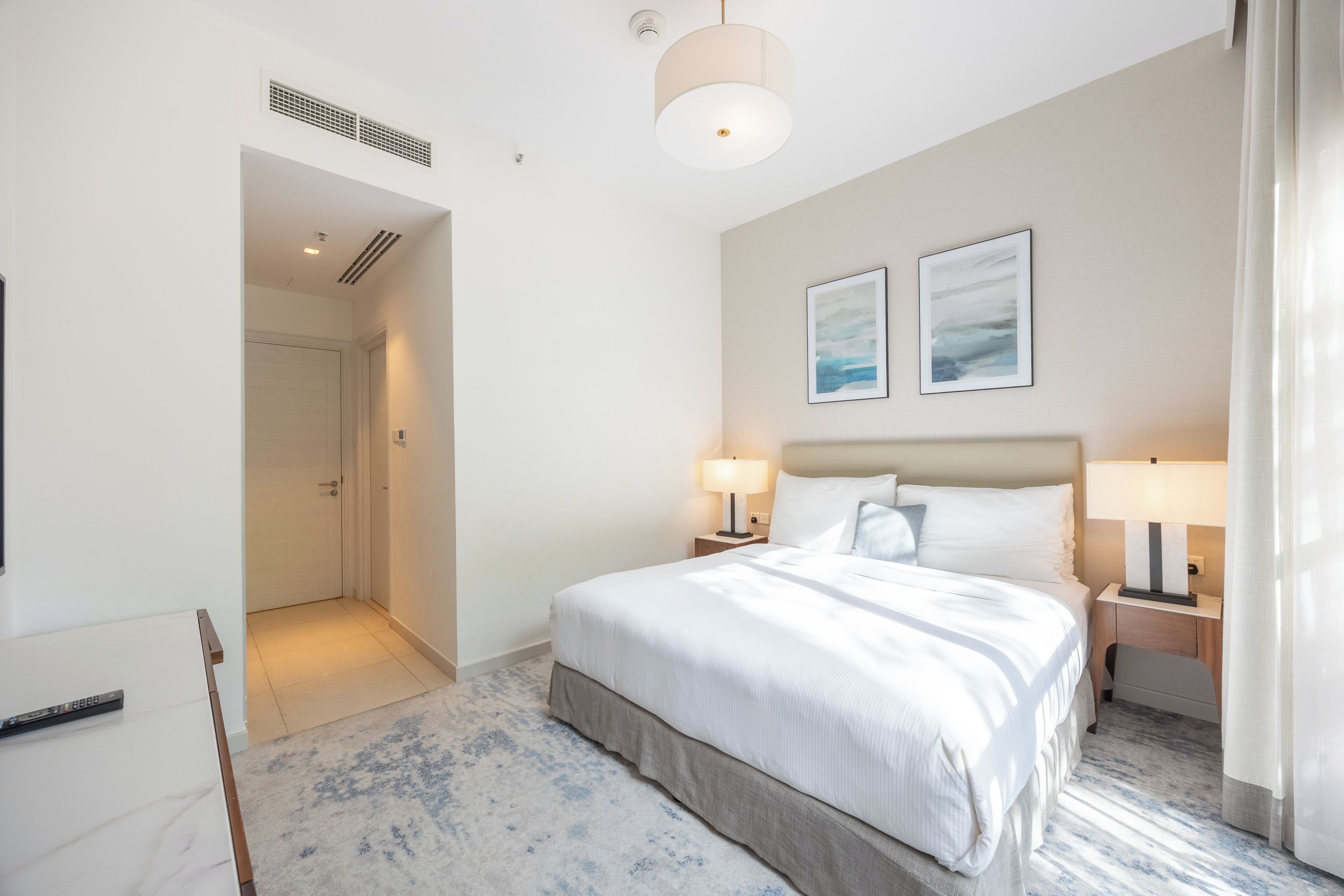 Een hotelkamer met een groot bed, twee bedlampen, een plafondlamp en een lijst met een abstract schilderij aan de muur.