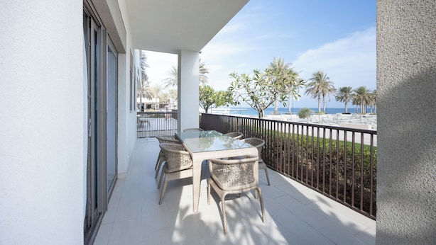 Een balkon met een tafel en stoelen, met uitzicht op een strand met palmbomen en een heldere lucht.