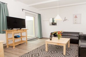 A living room with a television on a wooden stand, a coffee table with a vase of flowers, a black leather sofa, and a patterned rug. - Terraced house - 2 bedrooms - 55 sqm (Ostseebad Rerik)