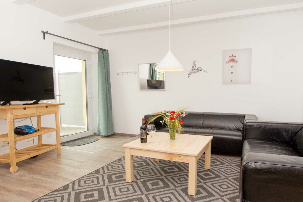 En stue med et fjernsyn på et træstel, et sofabord med en vase med blomster, en sort lædersofa og en gulvtæppe med et mønster.