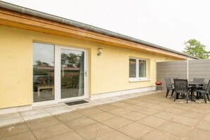 Ein Gebäude mit gelber Fassade, einer Schiebetür, einer kleinen Terrasse mit Tisch und Stühlen sowie einem grauen Zaun.