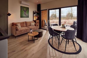 Un salon moderne avec un canapé, une table basse et un coin repas avec des chaises. Un tableau orne le mur et de grandes fenêtres offrent une vue sur l’extérieur.