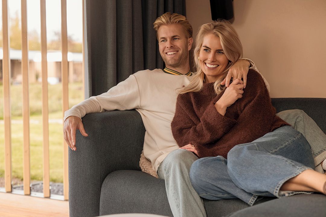 Een koppel zit op een grijze bank in een kamer met een groot raam en gordijnen.