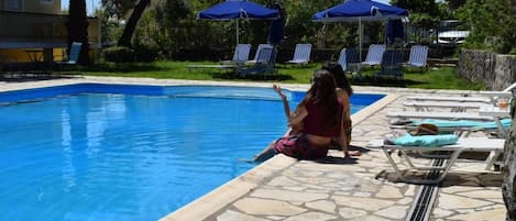 Piscina con sillas de descanso y sombrillas, rodeada de follaje y un edificio.