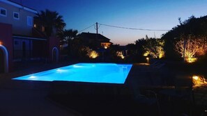 En swimmingpool i skymningen, omgiven av hus och med gatubelysning.