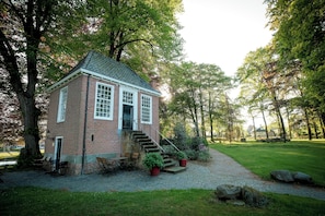 Un bâtiment en briques avec un escalier menant à une porte, entouré d’arbres et d’une zone pavée aménagée avec du mobilier d’extérieur.