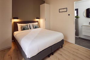Une chambre à coucher moderne, avec un lit, des lampes de chevet, une armoire et une salle de bain visible à travers une porte ouverte.