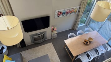 A modern living room with a flat-screen TV, a dining area with a wooden table and white chairs, and a large window with a view of greenery.