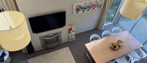 En moderne stue med et fladskærms-tv, et spiseområde med et træbord og hvide stole, samt et stort vindue med udsigt over grønne områder.