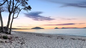 A beach with white sand, trees, and a calm sea with distant landforms. - Moola Haven – Hawks Nest (Hawks Nest)