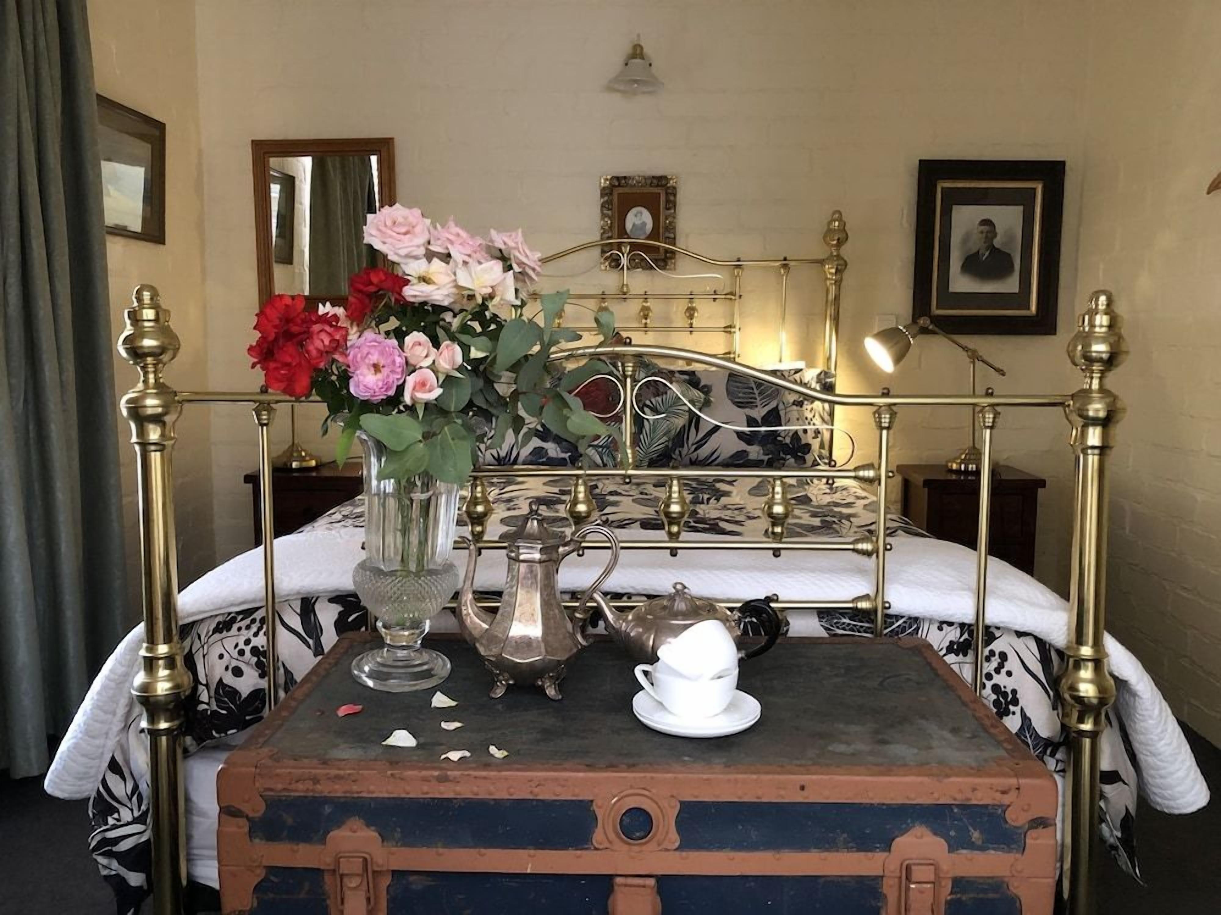 A bed with a metal frame, a vintage suitcase, a teapot, a cup, and a vase with flowers.