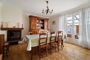 A dining room with a wooden table, chairs, a fireplace, and a large window with curtains. - Chez Monique: family house in Saint Coulomb near beaches (Saint-Coulomb)
