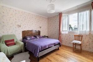 Ein Schlafzimmer mit floralem Wandpapier, einem hölzernen Bett, einem grünen Sessel, einem Holzstuhl und einem Fenster mit Vorhängen.